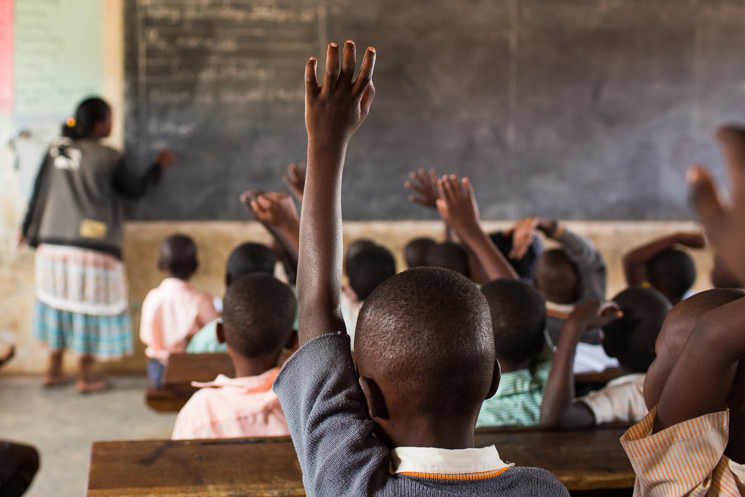 Children raising hands in a classroom in Uganda while the teacher writes on the board